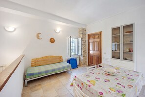 A room with a bed, a table with a fruit-patterned tablecloth, a cabinet with glass doors, and a stone wall.