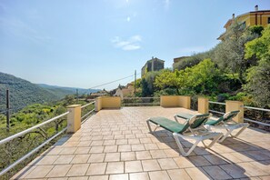 Eine Terrasse mit Blick auf eine hügelige Landschaft, ein Gebäude mit gelber Fassade und eine Steinmauer.