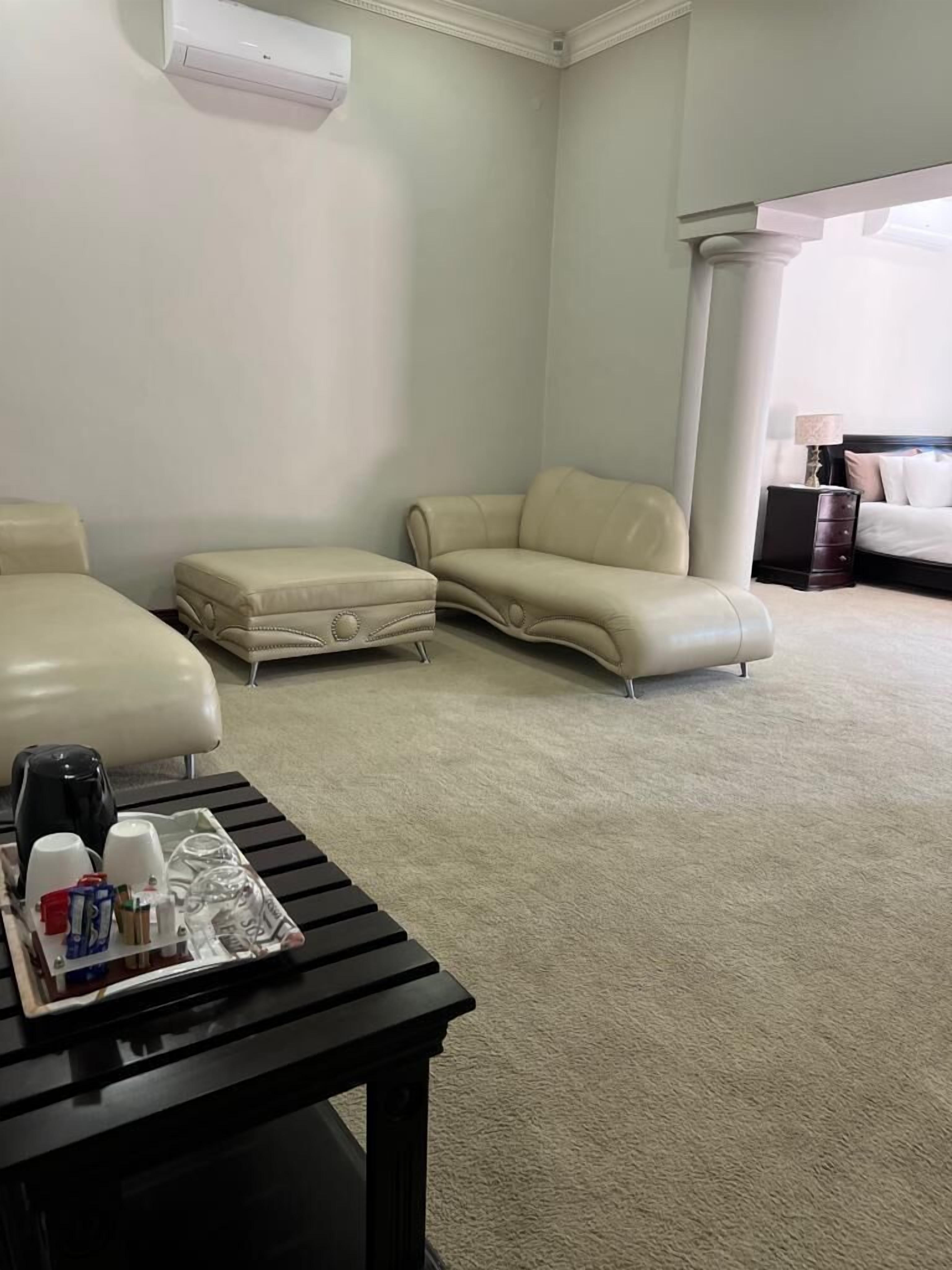 A room with a beige carpet, a black table with a tray of items, two beige sofas, a bed with a dark headboard, and a white air conditioning unit on the wall.