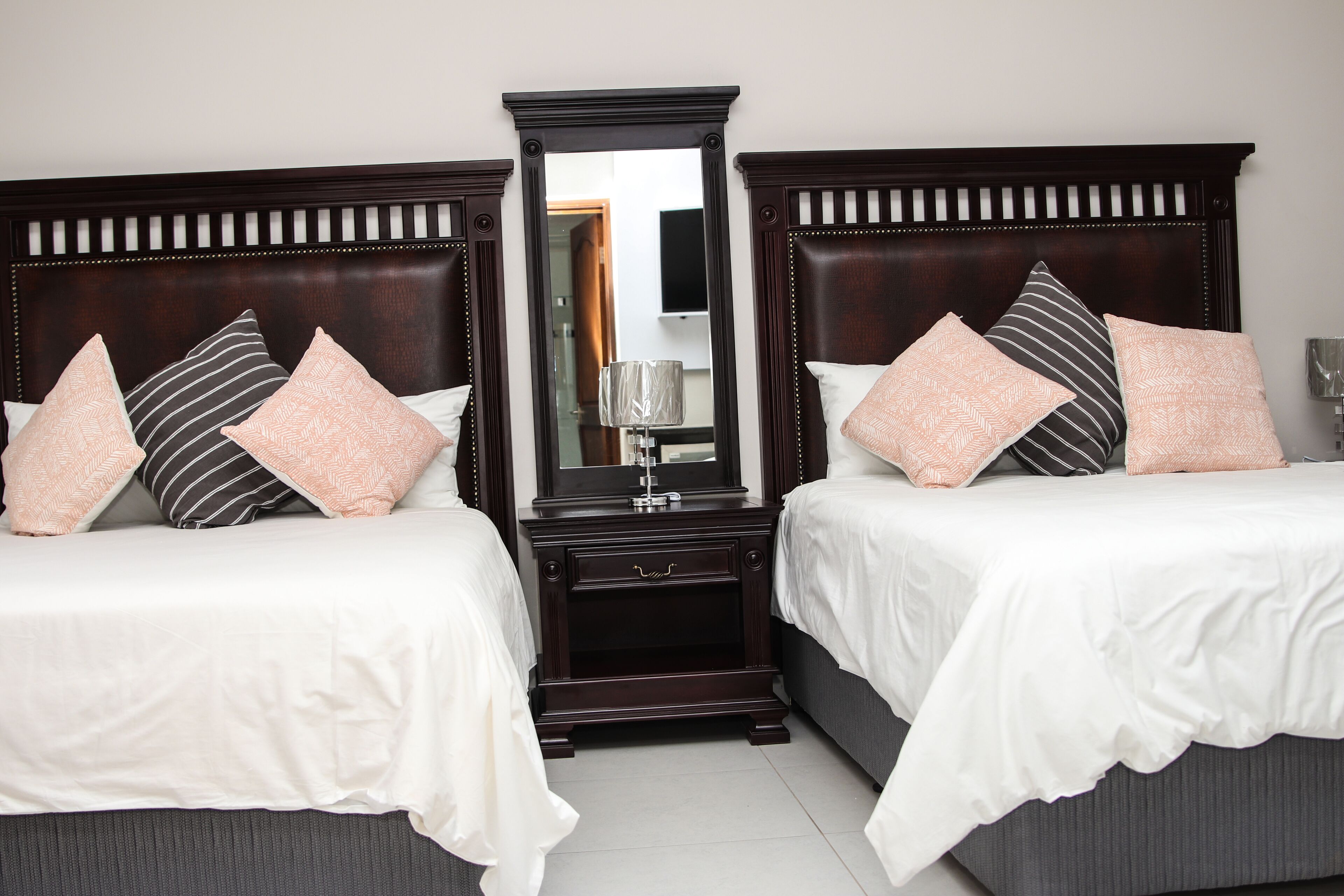 Une chambre d’hôtel avec deux lits, chacun doté d’une tête de lit en bois foncé, de draps blancs et de coussins décoratifs. Un grand miroir est placé au centre, entre les deux lits, et une petite table de chevet avec une lampe est visible.
