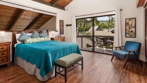 A bedroom with a large bed, a wooden ceiling, a balcony with a chair, and a view of palm trees.
