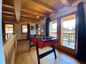 A room with a foosball table, wooden flooring, a balcony with a railing, and a view of a house outside.