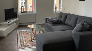 Ein modernes Wohnzimmer mit einem grauen Sofa, einem Glastisch, einem Flachbildfernseher auf einem weißen Standfuß und einem gemusterten Teppich.