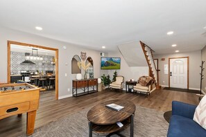 A spacious living area with a foosball table, a round coffee table, and a sofa. A staircase leads to an upper level.