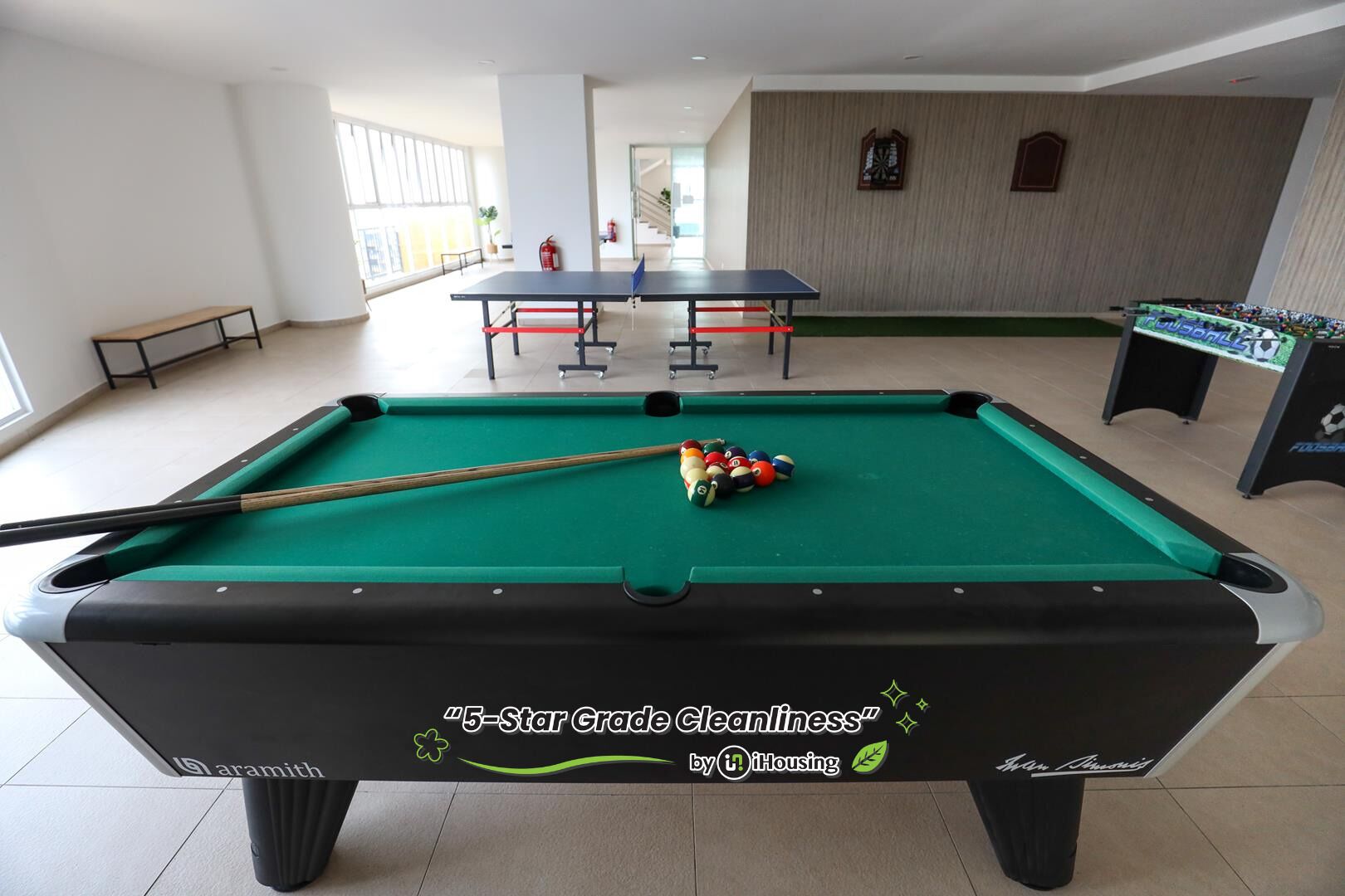 Un tavolo da biliardo con stecca e palle, un tavolo da calcio e una panca in un ambiente interno ben illuminato.