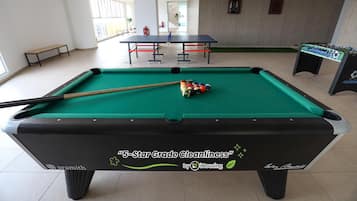 Un tavolo da biliardo con stecca e palle, un tavolo da calcio e una panca in un ambiente interno ben illuminato.
