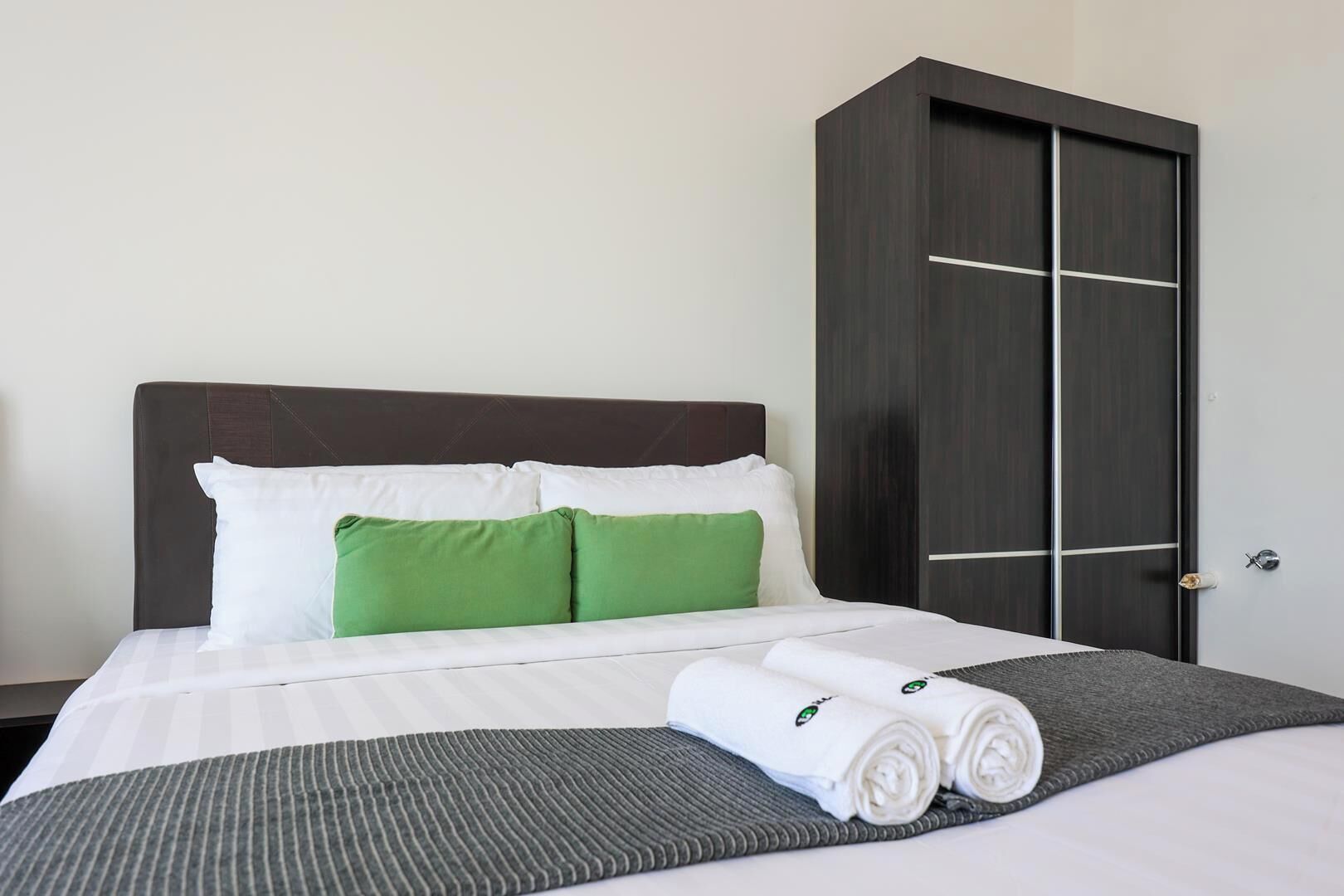 A hotel room with a bed, pillows, a dark wardrobe, and a rolled towel on the bed.