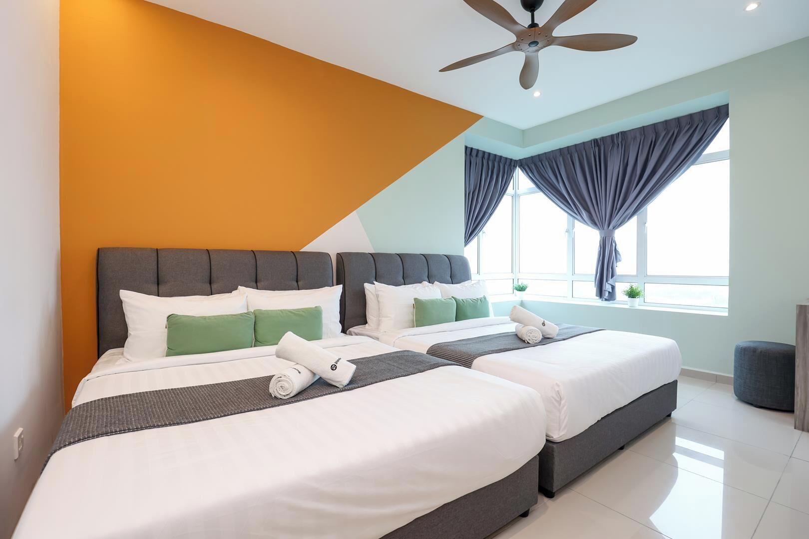 Quarto de hotel com duas camas, ventoinha no teto, janela grande com cortinas e vista para o mar.