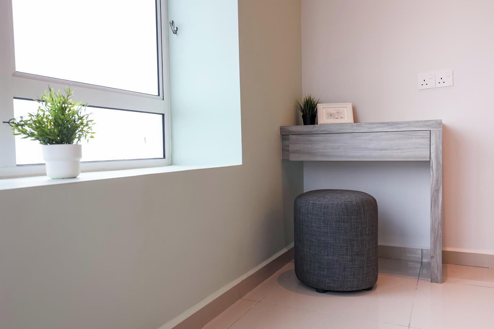 Una habitación con ventana, una planta en maceta, un reposapiés gris y una estantería con un marco de fotos.