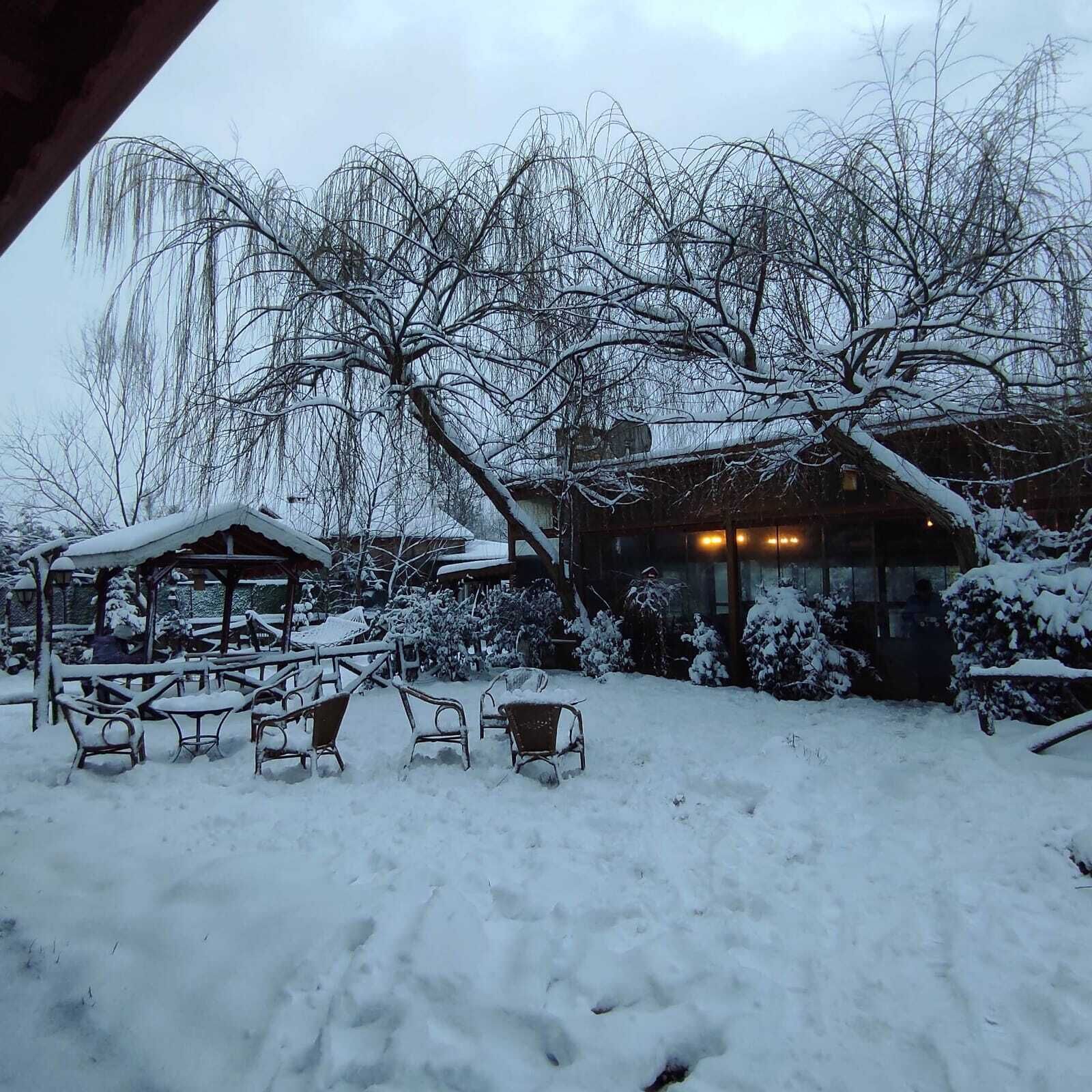 A snow-covered outdoor seating area with tables and chairs, a gazebo, and a building with illuminated windows.