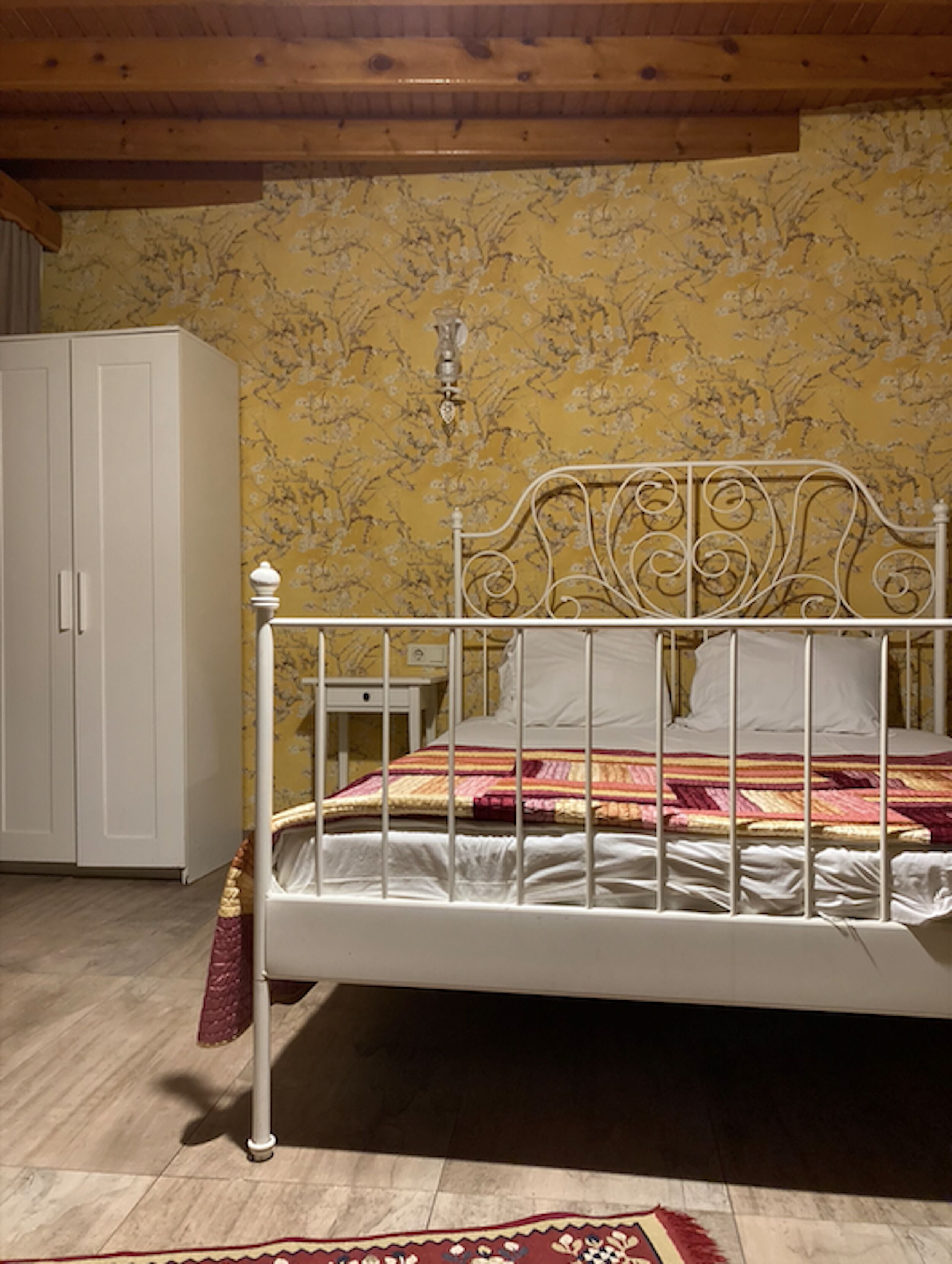 A bedroom with a white metal bed frame, a wooden wardrobe, and a patterned wallpaper.