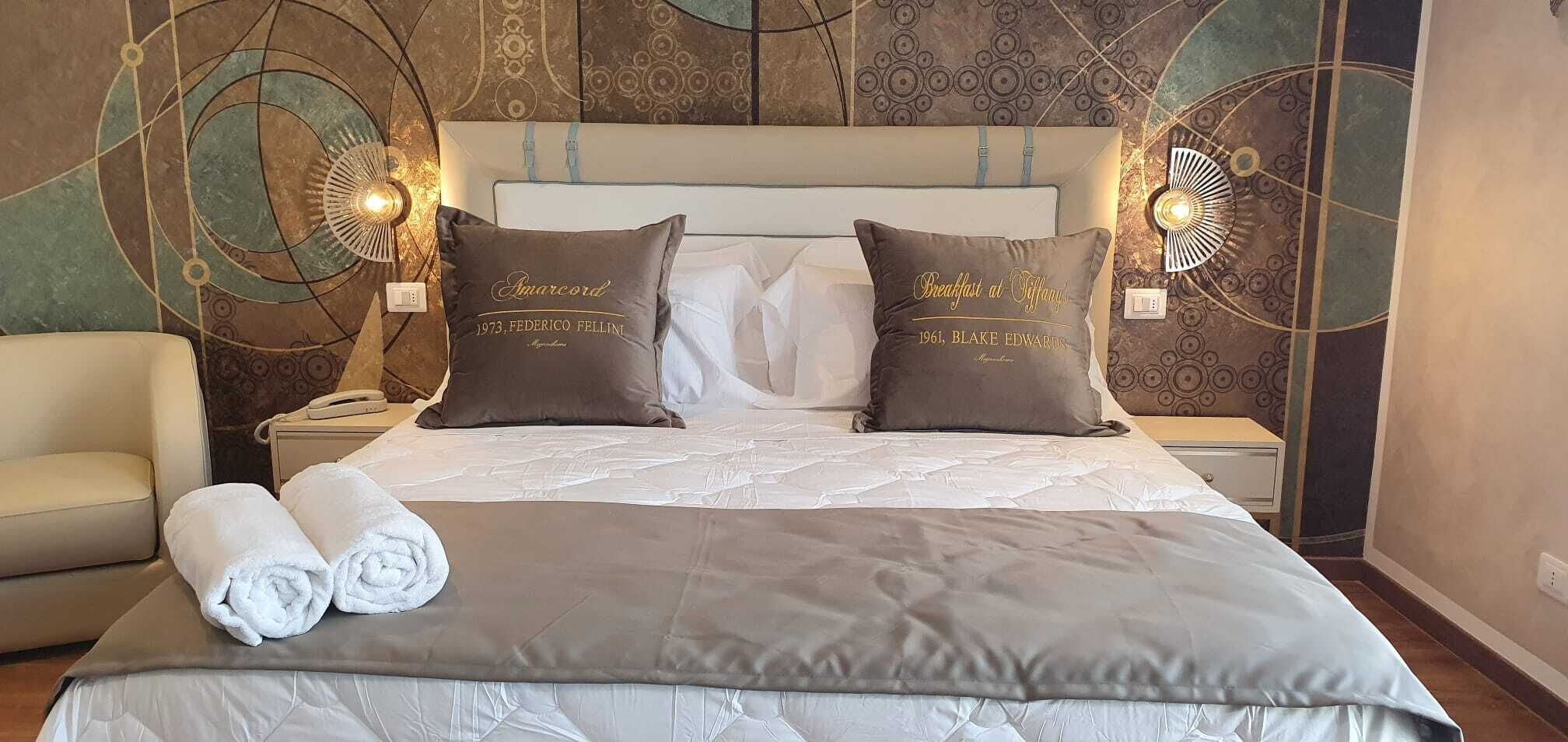 A neatly made bed with two decorative pillows, a pair of rolled towels, and a headboard with a light fixture.