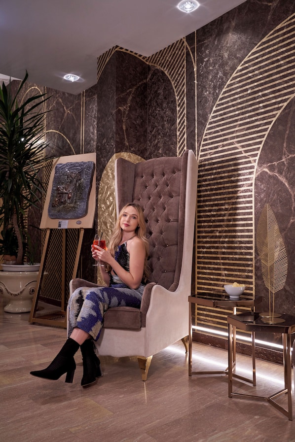 A woman seated in a tufted armchair with a painting on an easel behind her, a small table with a bowl, and a potted plant to the left. - Amsterdam Suite Hotel & SPA (Rimini)