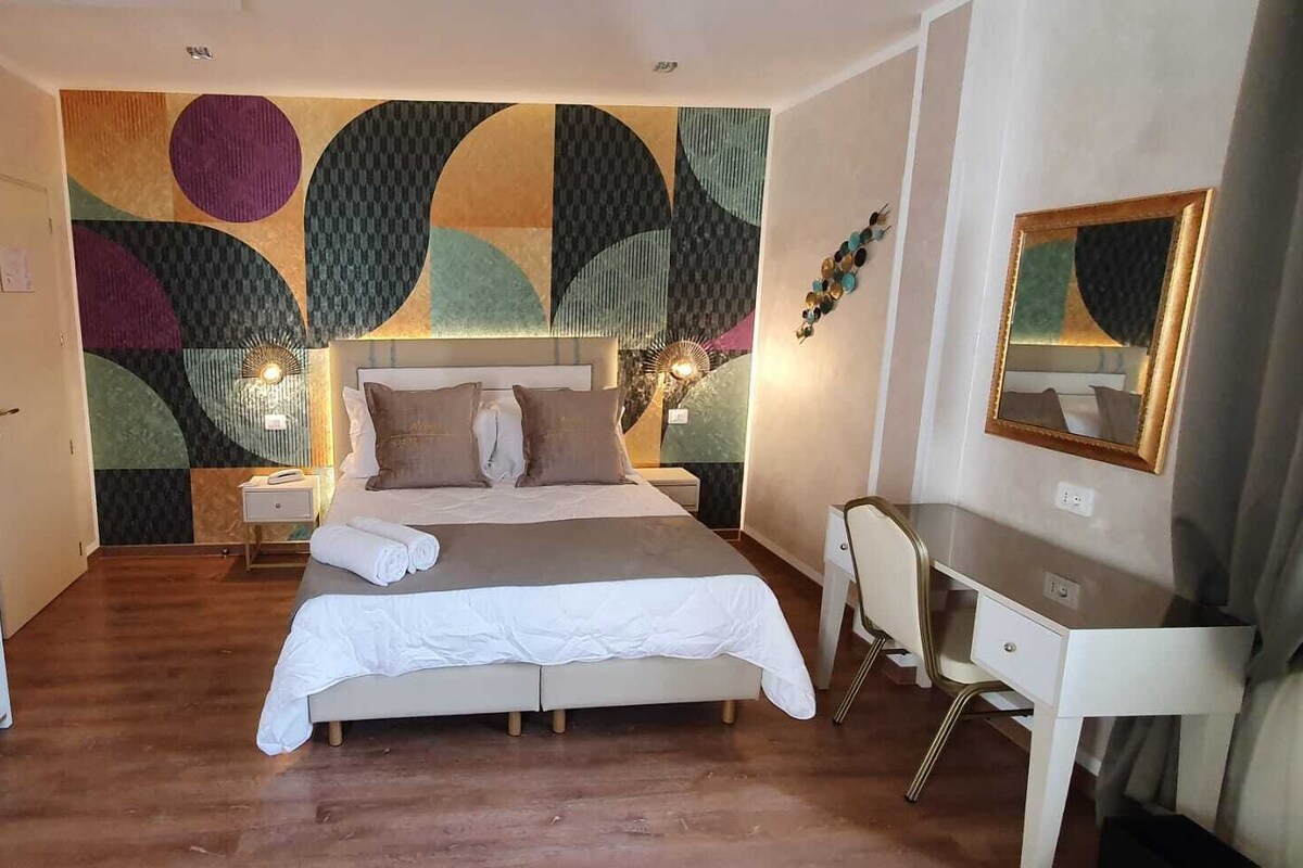 A hotel room with a large bed, a desk, and a chair. There is a wardrobe and a mirror on the wall.