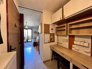 Corredor que dá para um quarto com cama e poltrona, área de cozinha com armários e quadro de avisos, e um quadro na parede.