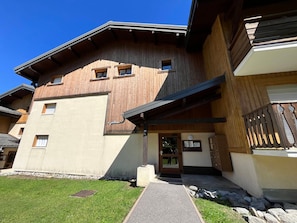 A multi-story wooden building with a flat roof, a visible entrance, and a small outdoor area with a pathway leading to the entrance.
