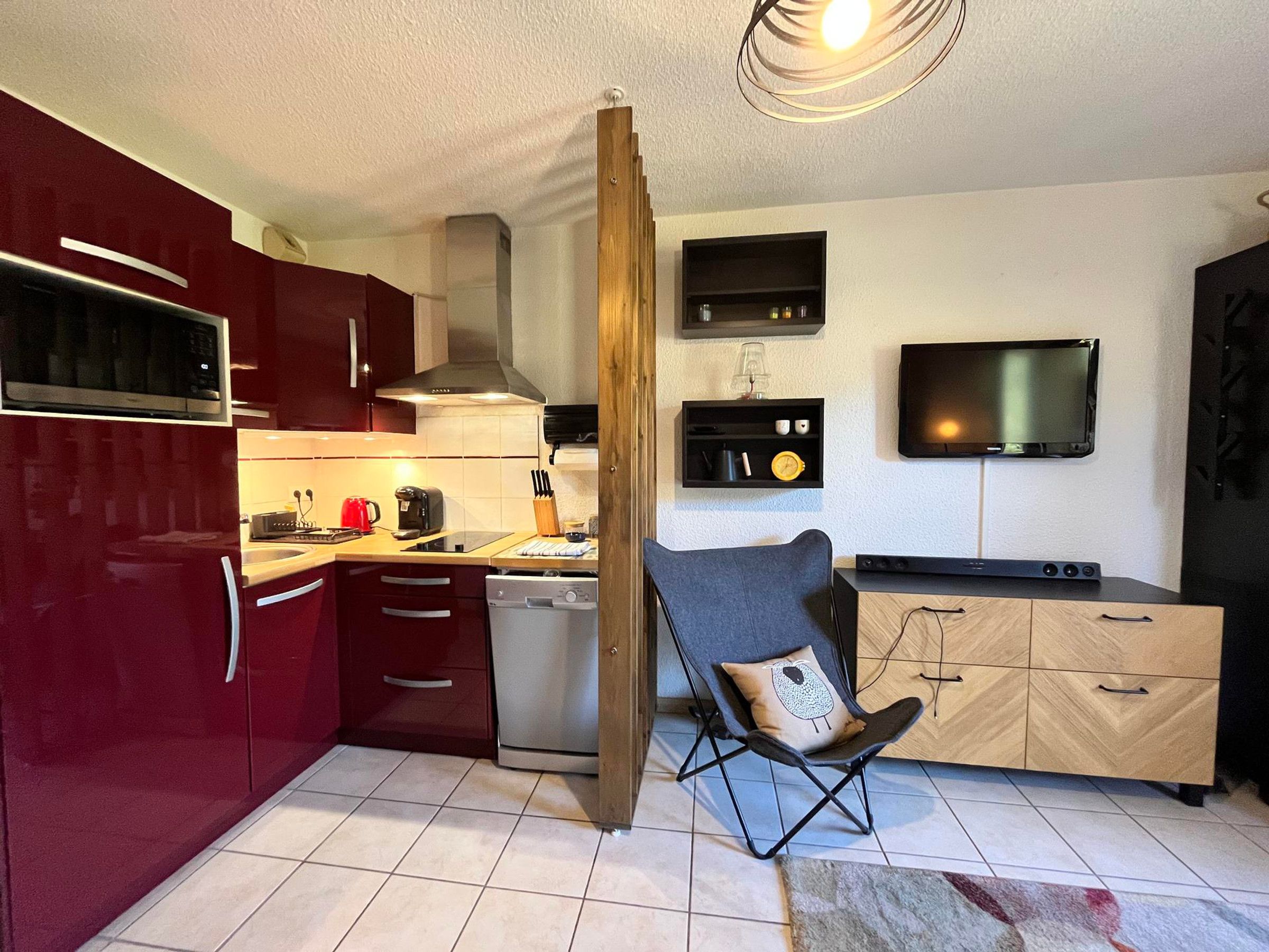 A compact kitchen with a microwave, stove, and sink. There's a TV mounted on the wall and a folding chair with a cushion.