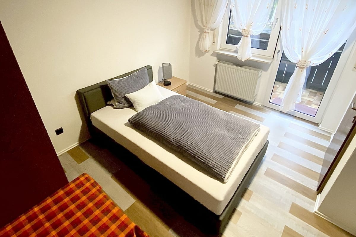 A bedroom with a bed, a nightstand, a radiator, and a window with curtains.