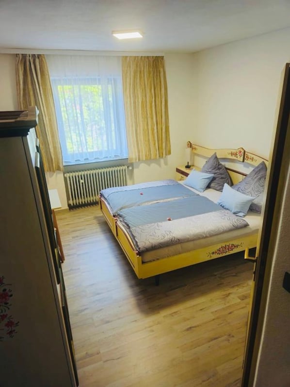 Ein Schlafzimmer mit einem Doppelbett, einem Heizkörper, einem Fenster mit Vorhängen und Holzdielen.