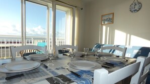 A dining table set for four with white chairs, a view of the sea through large windows, and a clock on the wall. - Sea view apartment in Notre-Dame-de-Monts - 5 people, pets (Notre-Dame-de-Monts)