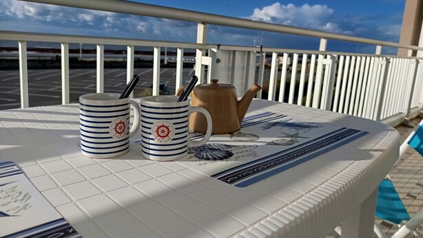 A table with a nautical-themed tablecloth, two striped mugs, and a teapot on a balcony with a railing and a view of a parking lot and sky. - Sea view apartment in Notre-Dame-de-Monts - 5 people, pets (Notre-Dame-de-Monts)