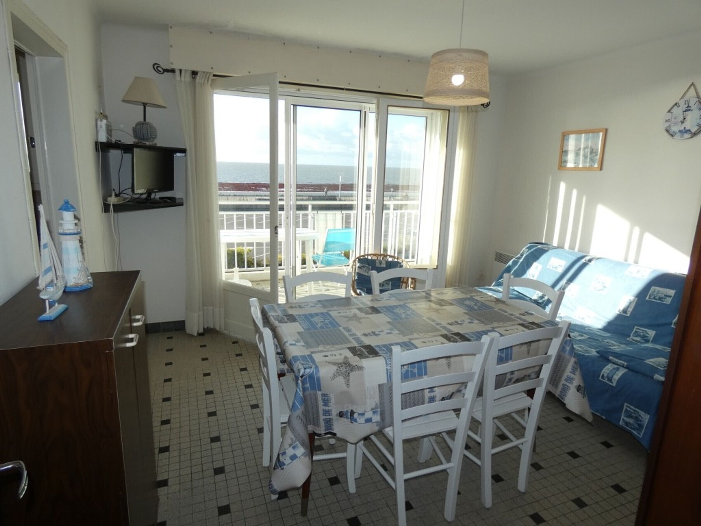Een kamer met een eettafel, stoelen, een kleine televisie en uitzicht op de zee door grote ramen.