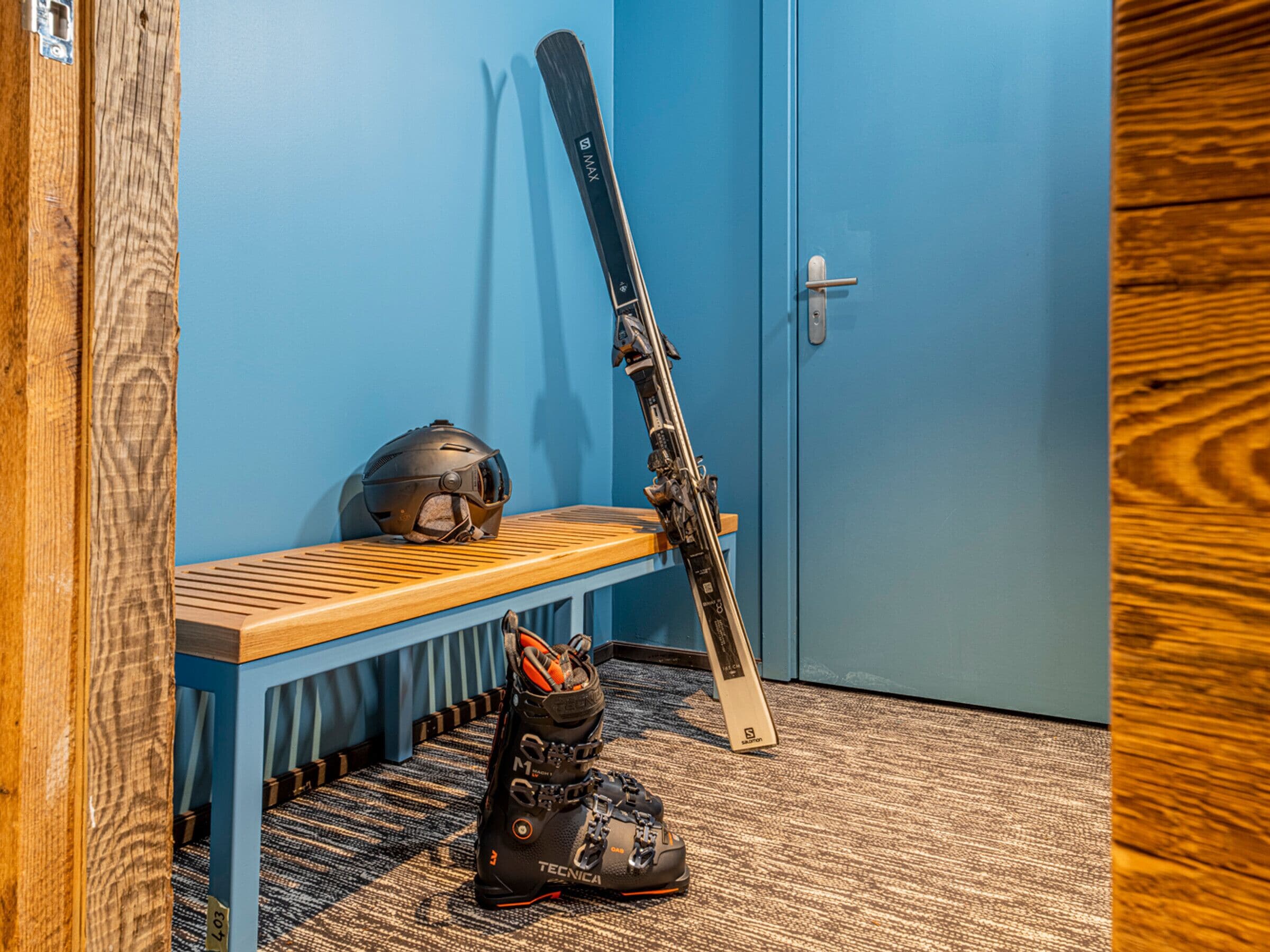 A room with a blue door, a wooden bench, a pair of skis leaning against the wall, and a pair of boots on the floor.