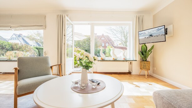 Una habitación luminosa con un ventanal grande, una mesa blanca, un sillón gris y una planta en maceta.