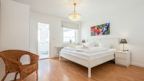 Un dormitorio con cama blanca, mesitas de noche, una silla de mimbre, ventana con persianas, radiador y lámpara en el techo.