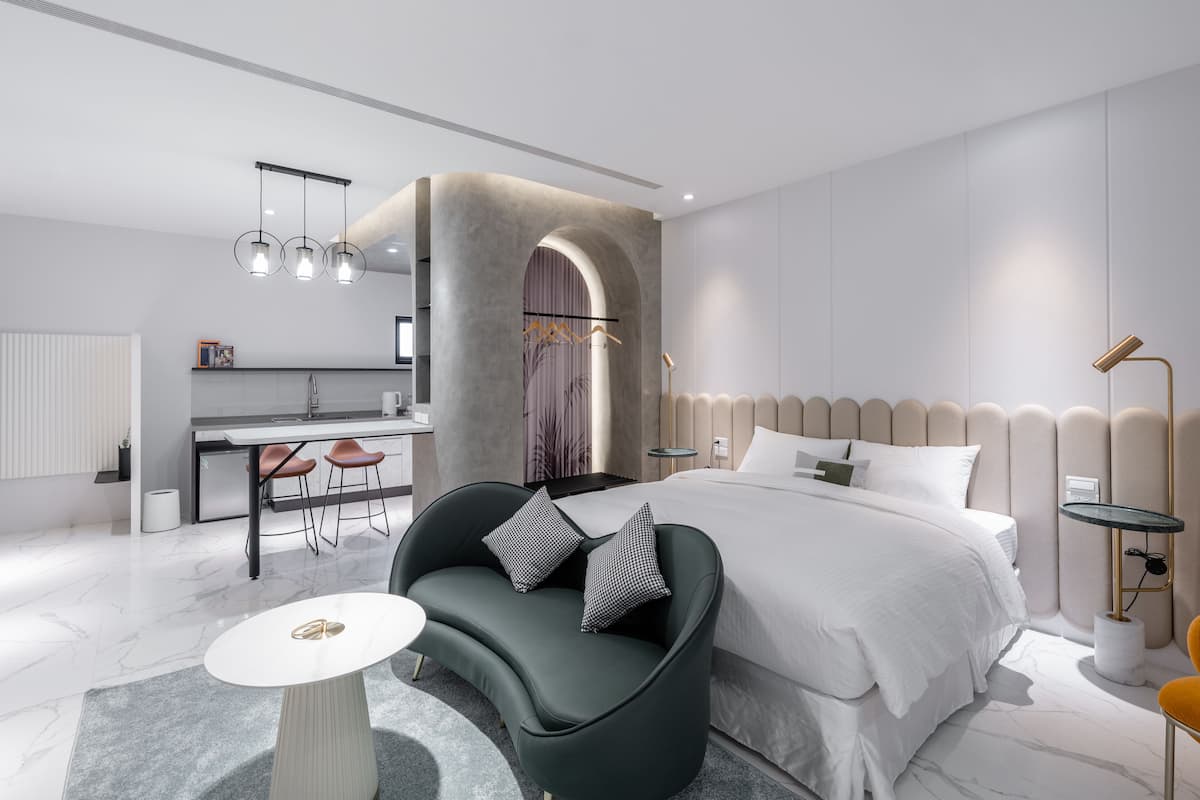 Un dormitorio moderno con una cama grande, un sofá curvo, una mesita redonda y una kitchenette al fondo.