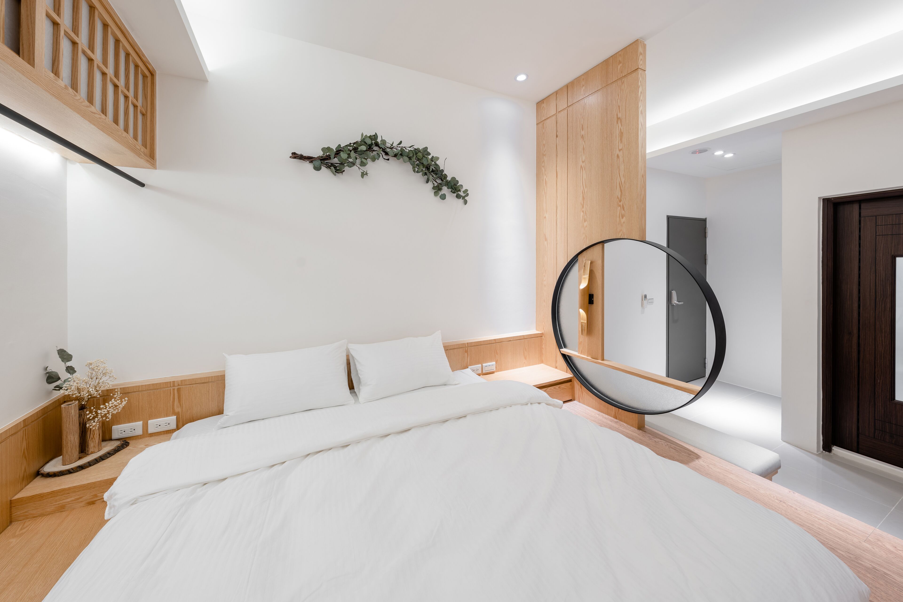 Un dormitorio moderno con una cama grande, cabecera de madera, un espejo circular y una mesita de noche con un jarrón de flores.