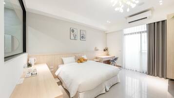 A modern hotel room with a large bed, a desk, and a chair. There is a window with curtains, an air conditioning unit, and artwork on the wall.