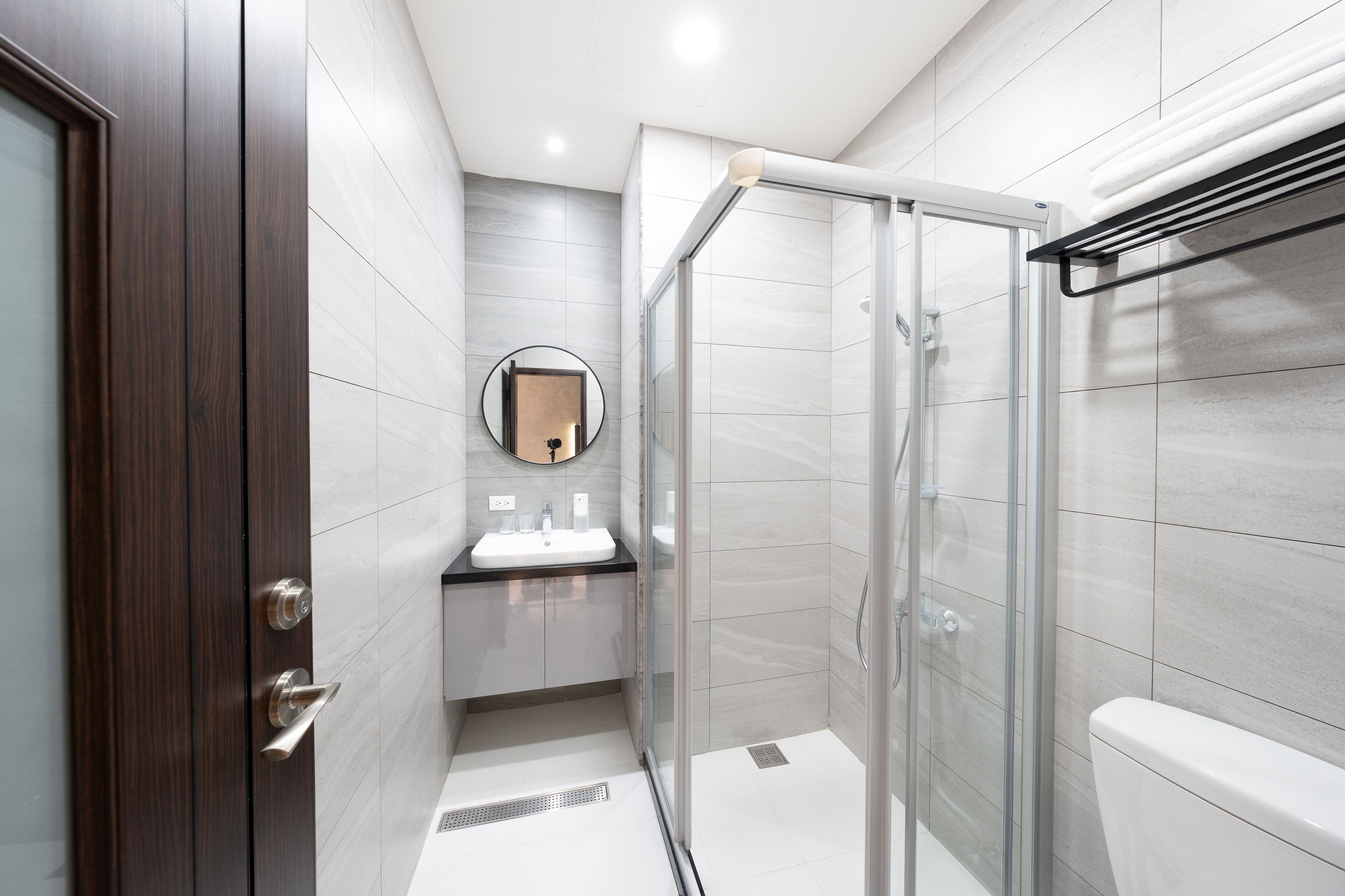 Un baño moderno con cabina de ducha de vidrio, un espejo redondo y una puerta de madera oscura.
