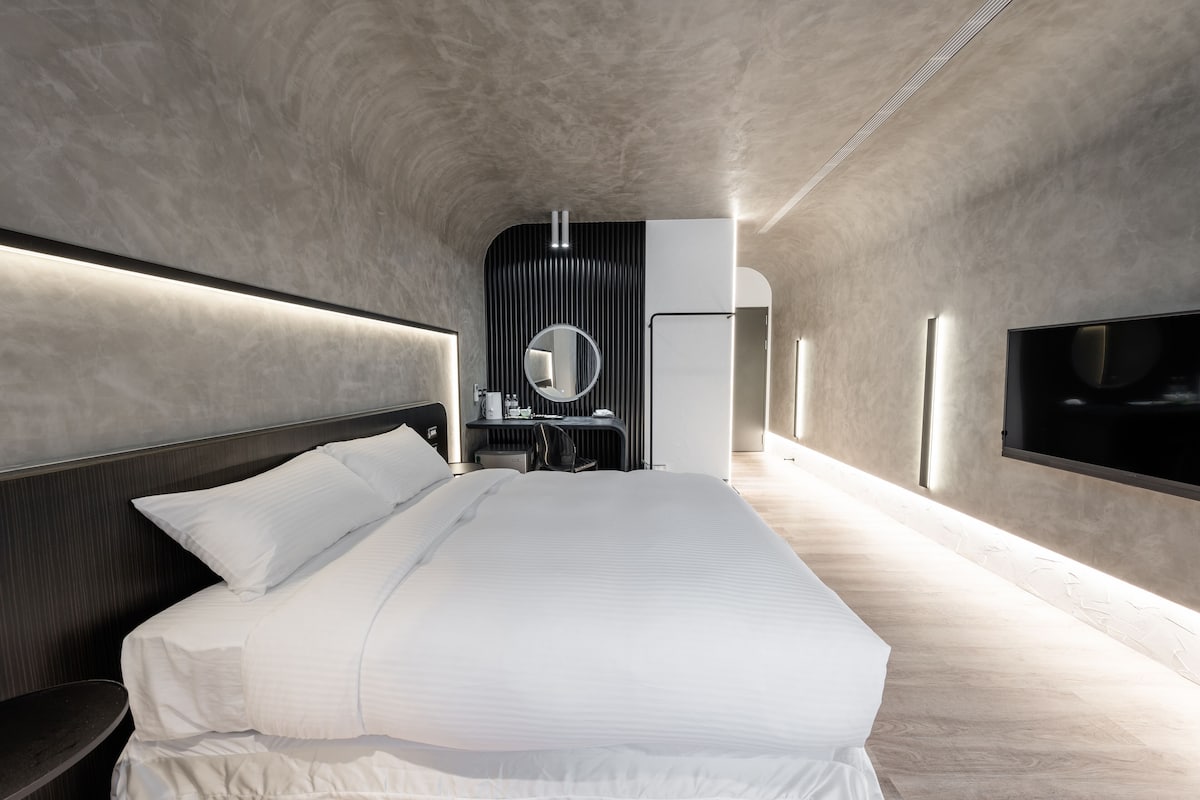 Un dormitorio moderno con una cama grande, una mesita de noche, un espejo, un escritorio y un televisor fijado a la pared.