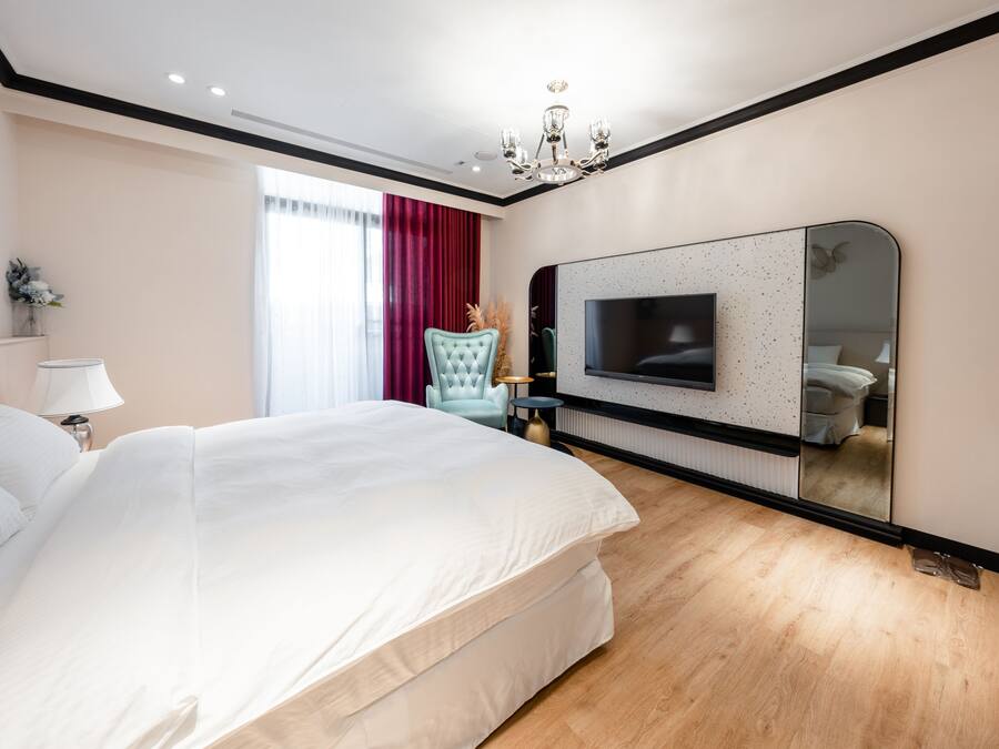 A modern bedroom with a large bed, a TV mounted on the wall, a chandelier, and a window with red curtains.