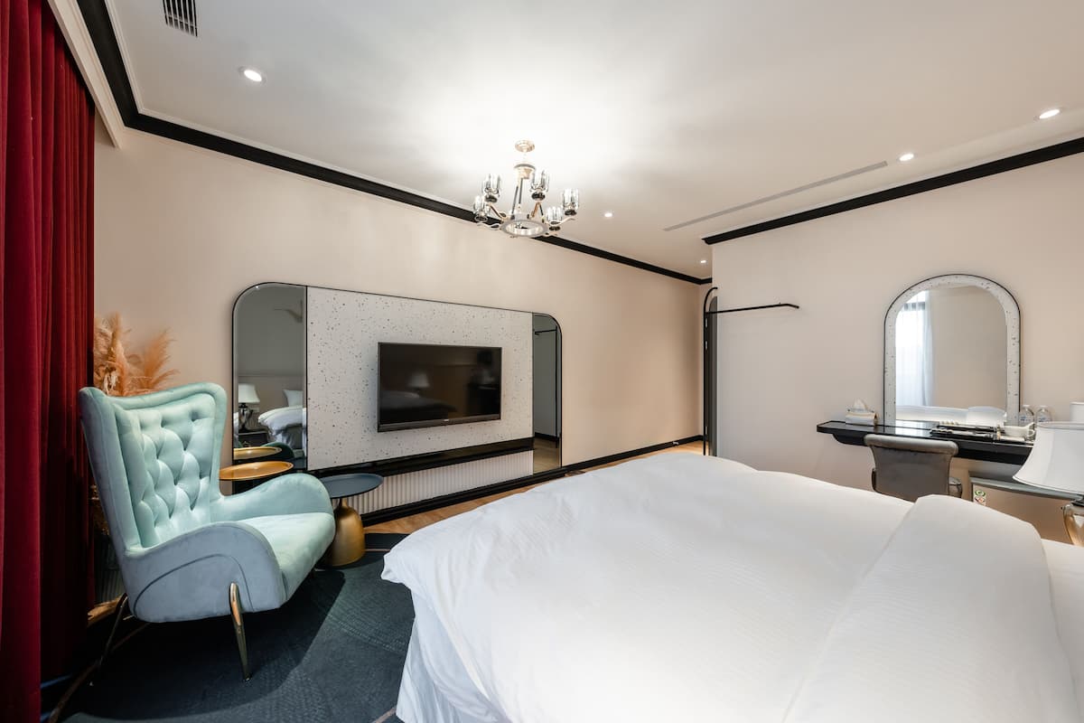 Una habitación de hotel moderna con una cama grande, un área de descanso con un sillón, un televisor montado en la pared, un espejo y una araña.