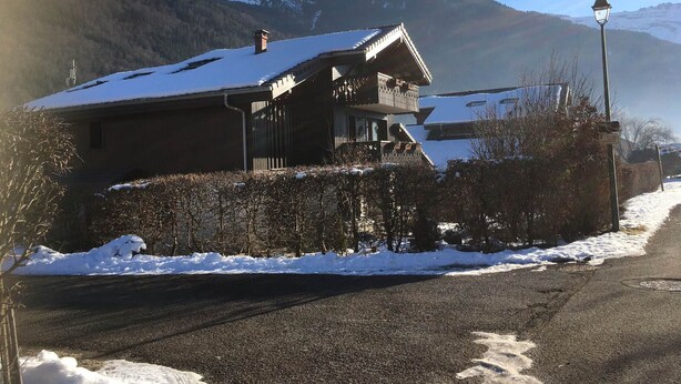 A two-story chalet with a snow-covered roof, situated by a road with a mountain backdrop. - Cozy apartment, 3 rooms, garden, quiet, near Samoëns center (Samoëns)