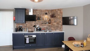 A modern kitchen with dark cabinets, a brick backsplash, and a flat-screen TV mounted on the wall. - Cathedral Superior Apartment (Worcestershire)