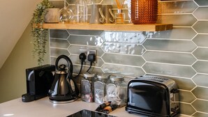 A kitchen with a toaster, kettle, and coffee maker, along with jars and a shelf with glassware and a plant.