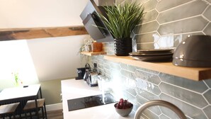A modern kitchen with a hexagonal tile backsplash, a wooden countertop, a black range hood, a potted plant, a bowl of grapes, and a dining area with a table and chairs.