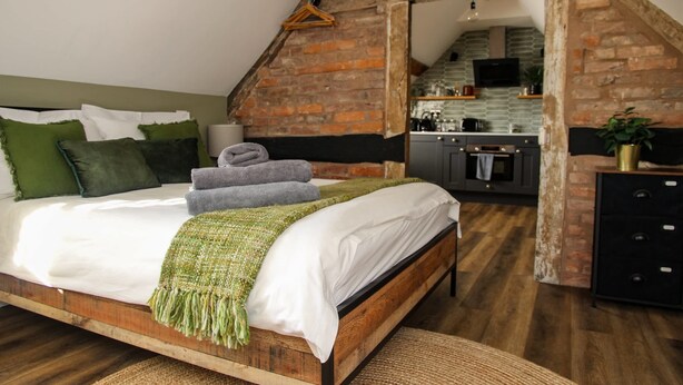 A bedroom with a wooden bed, green and white pillows, a green throw blanket, and a kitchen in the background.