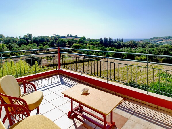 A balcony with a table and chairs, offering a view of a green landscape and the sea.