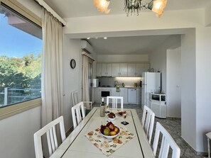A dining area with a table set for four, a kitchen in the background, and a view of greenery outside.