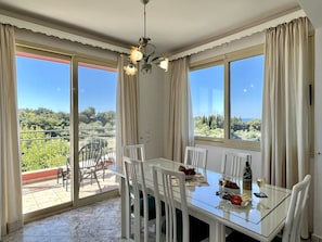 A dining area with a glass table, white chairs, a chandelier, and a view of greenery and trees through large windows.