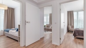 A modern hotel room with a wooden floor, a bed with a grey headboard, a bedside table, and a chair. There is a door leading to another room.