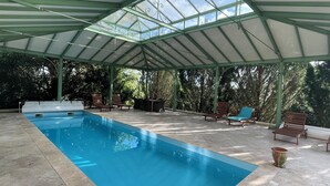 Una piscina interior con techo de cristal, rodeada de sillones de descanso y un porche cubierto.
