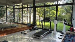 Un gimnasio con equipamiento de ejercicio, una mesa de fútbol de mesa y amplias ventanas con vistas a zonas verdes.