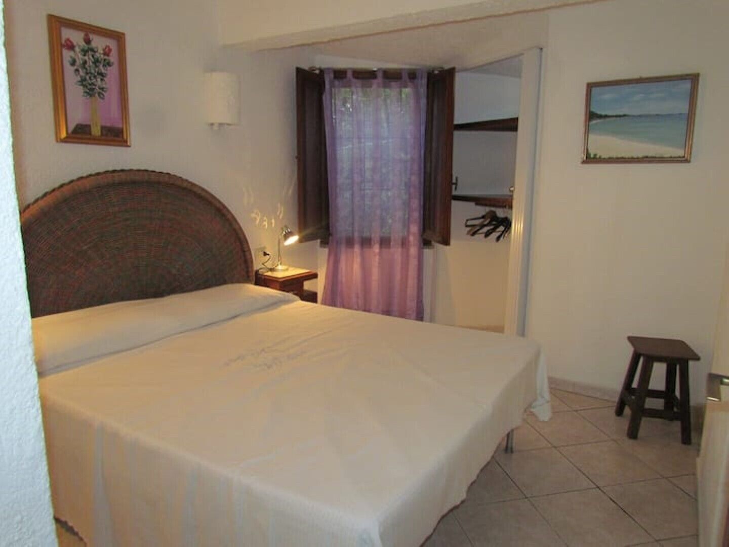 A bedroom with a bed, a nightstand, a lamp, a small stool, and a painting on the wall.