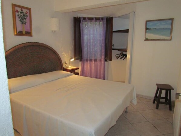 A bedroom with a bed, a nightstand, a lamp, a small stool, and a painting on the wall.