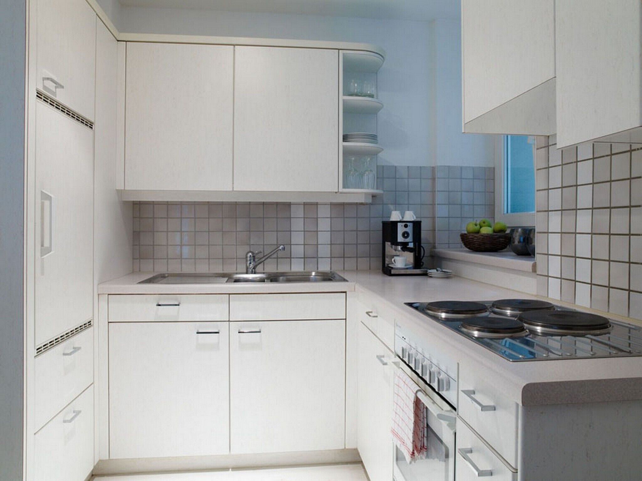 A modern kitchen with white cabinetry, a built-in stove, a coffee maker, and a window with a view.